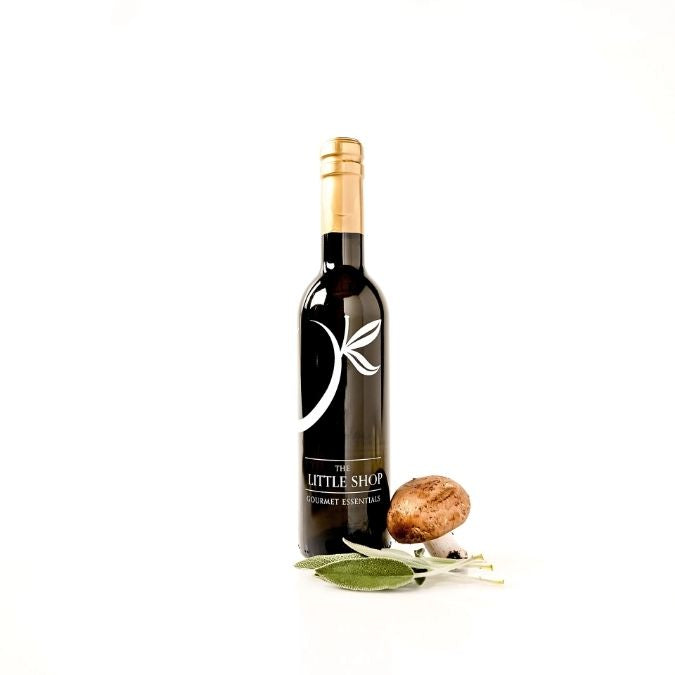 A bottle of gourmet olive oil from 'The Little Shop' with a gold foil cap, styled with sage leaves and a mushroom on a clean white background.