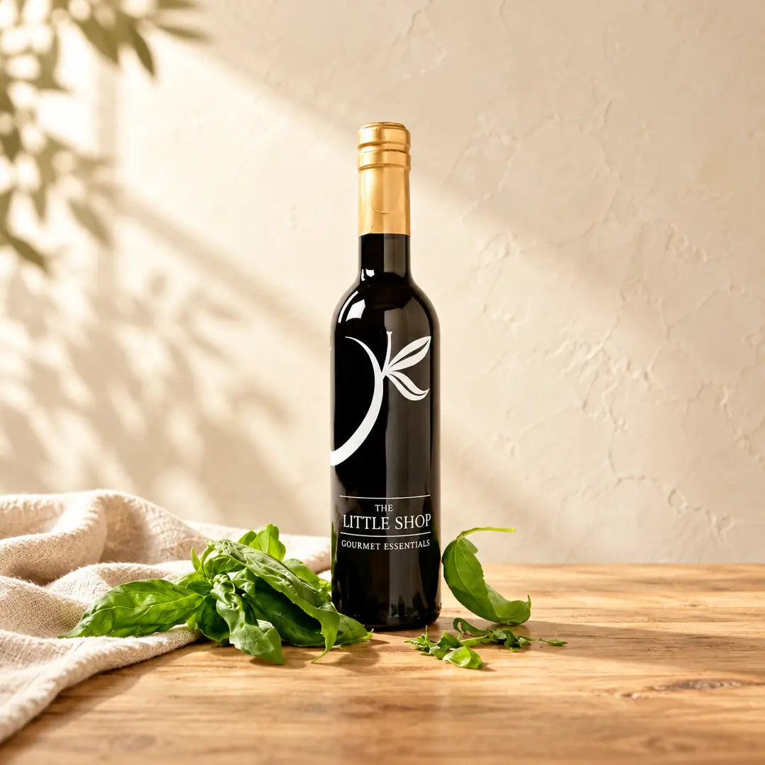 A dark glass bottle of Basil Premium Olive Oil with a gold top, displayed alongside fresh green basil leaves on a light wooden surface with soft leaf shadows.