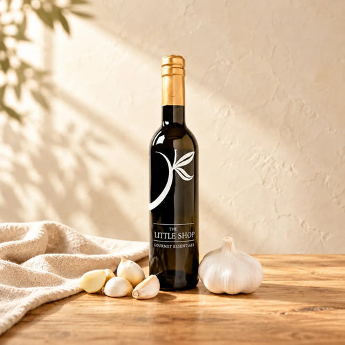 A dark glass bottle of Garlic Premium Olive Oil with a gold foil top and a white leaf logo, standing on a light wooden table next to a whole garlic bulb, peeled cloves, and a folded linen cloth under soft leaf shadows at The Little Shop of Olive Oils.