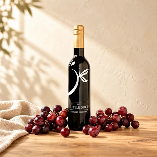A dark glass bottle of Traditional Dark Balsamic Vinegar with a gold foil top and a white leaf logo, standing on a light wooden table next to a cluster of fresh dark red grapes and a folded linen cloth under soft leaf shadows at The Little Shop of Olive Oils.