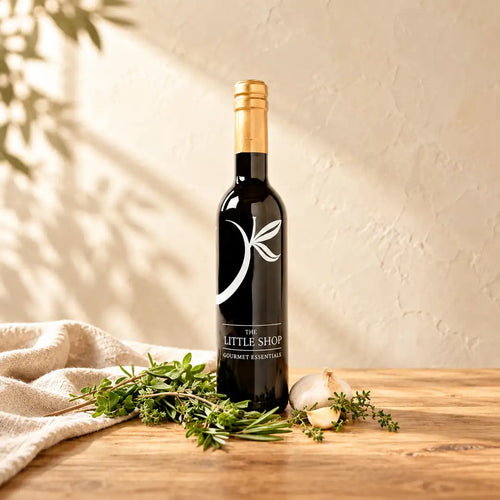 A dark glass bottle of Tuscan Herb Premium Olive Oil with a gold foil top and a white leaf logo, standing on a light wooden table next to fresh sprigs of rosemary, thyme, and garlic cloves, with a folded linen cloth under soft leaf shadows at The Little Shop of Olive Oils.